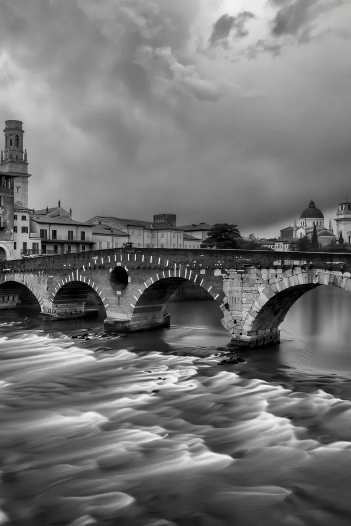 Ponte Pietra, il ponte romano di Verona, in bianco e nero: l'Adige impetuoso sotto le arcate e il centro storico sotto un cielo di tempesta